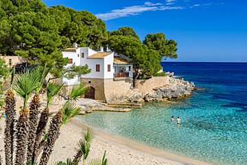 A picturesque cove with white houses nestled among pine trees, a sandy beach, and clear turquoise waters where two people swim under a bright blue sky.
