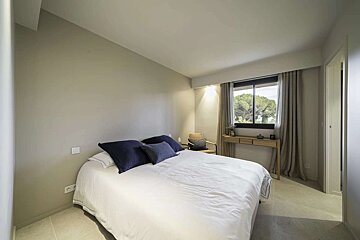 A serene, modern bedroom features a white bed with blue pillows, a large window overlooking trees, and neutral-toned walls. A small wooden console table and woven chair complete the minimalist space.