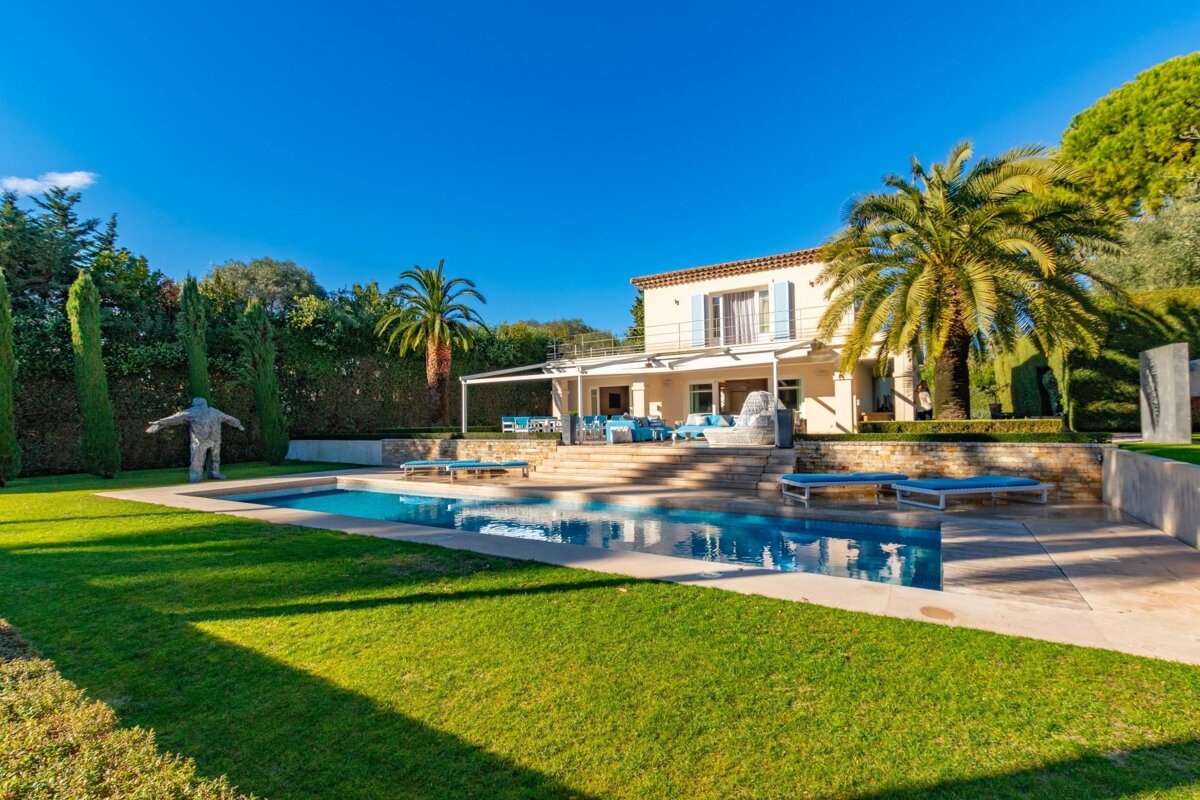 A luxurious two-story villa with a large swimming pool, green lawn, palm trees, and loungers under a clear blue sky.