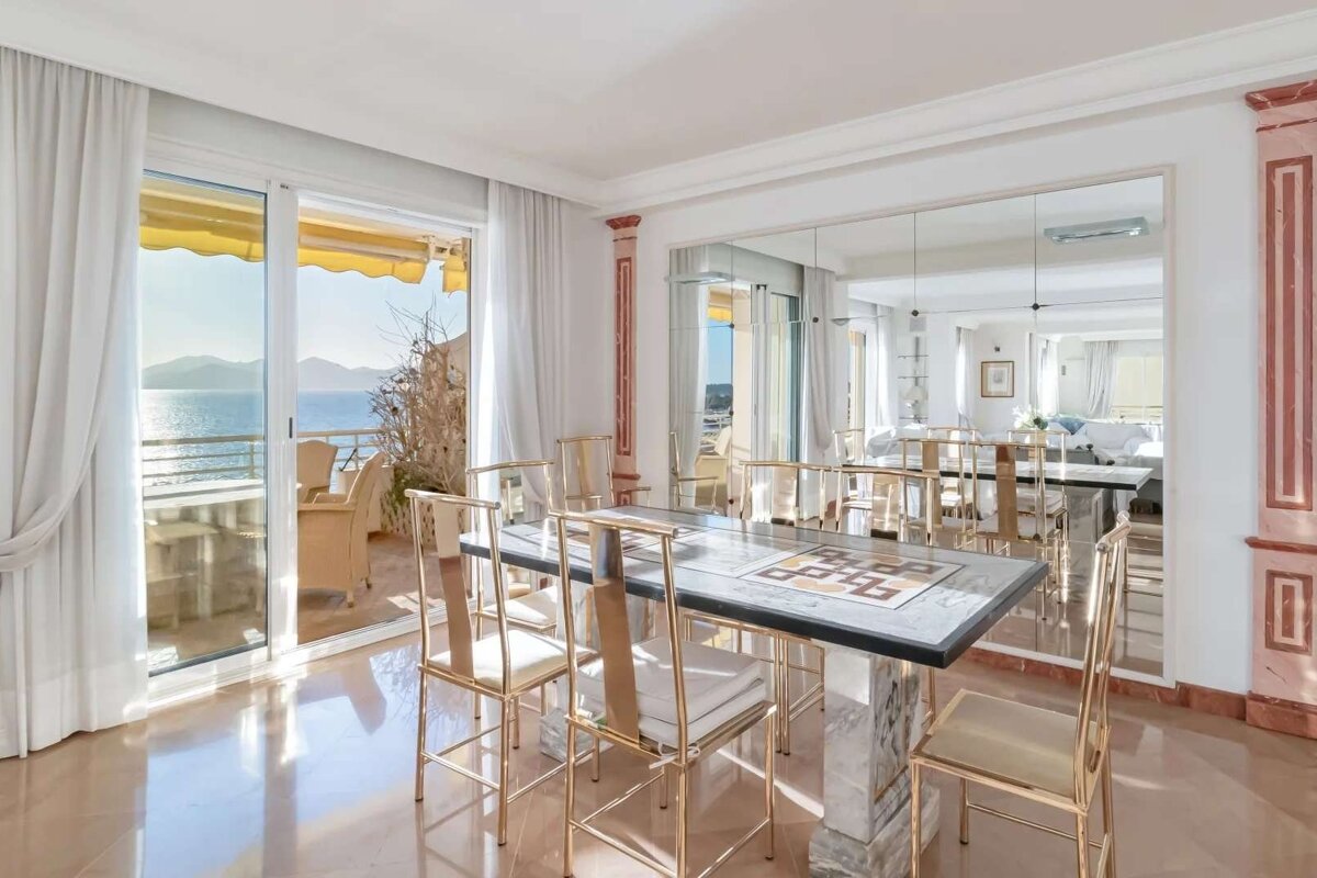 Sunny dining room with gold chairs, stone table, and large sliding doors opening to a balcony with a beautiful ocean and mountain view.