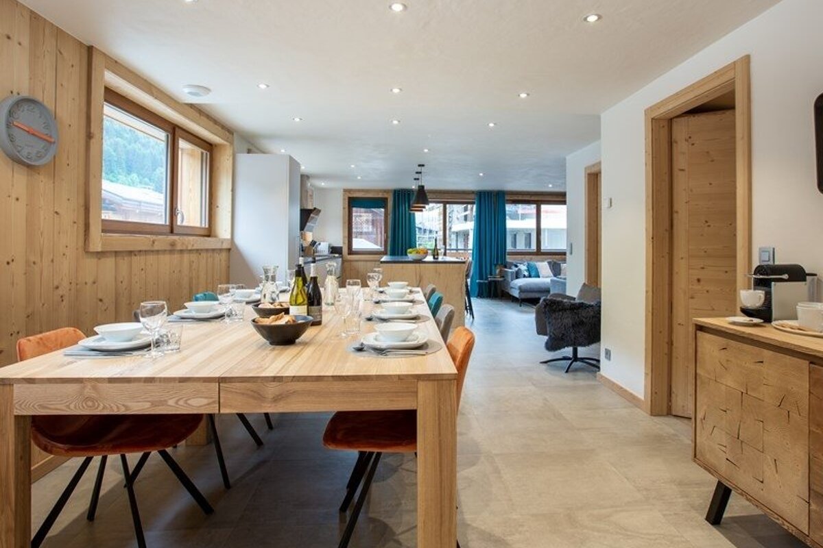 A modern chalet dining area with a long wooden table set for a meal, opening into a living space with wooden paneling.