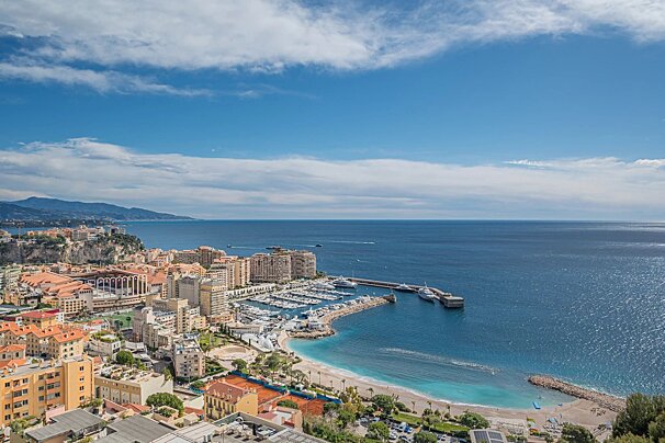 A sunny coastal view of Monaco showing a yacht harbor, sandy beach, cityscape, and the deep blue Mediterranean Sea.