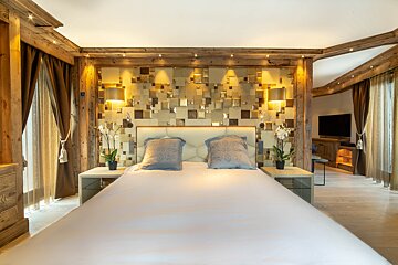 A luxurious bedroom features a large white bed, an ornate square-patterned wall behind the headboard, and wooden chalet-style accents. A TV is visible.