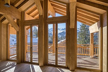Bright, timber-framed room with large glass doors opening to a balcony with a panoramic view of sunny, snowy mountains and pine trees.