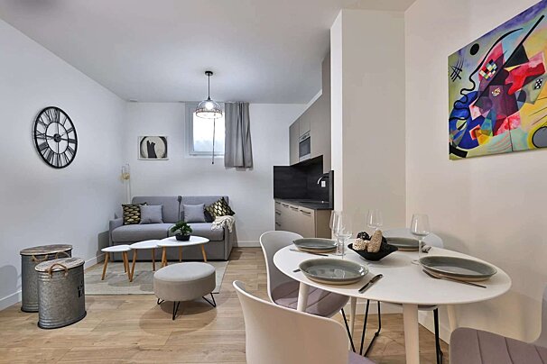 A modern, open-plan studio apartment featuring living, dining, and compact kitchen areas. Neutral decor, abstract art, a large clock, and a stylish gray sofa.