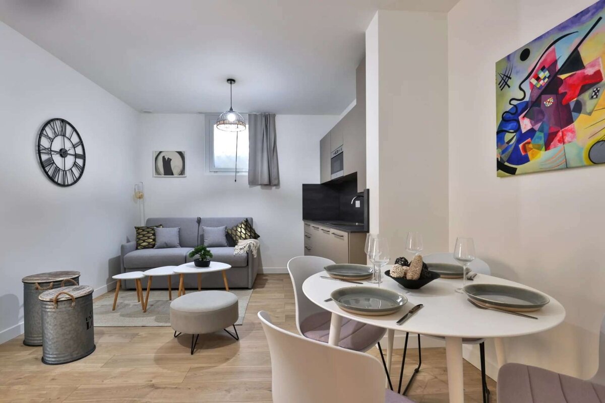 A modern, open-concept living space with a gray sofa, white dining table, and a colorful abstract painting. Features wood floors and a compact kitchen.