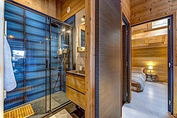Modern bathroom with a blue tiled shower, wooden vanity, and a glimpse into a cozy, wood-paneled bedroom through an open doorway.