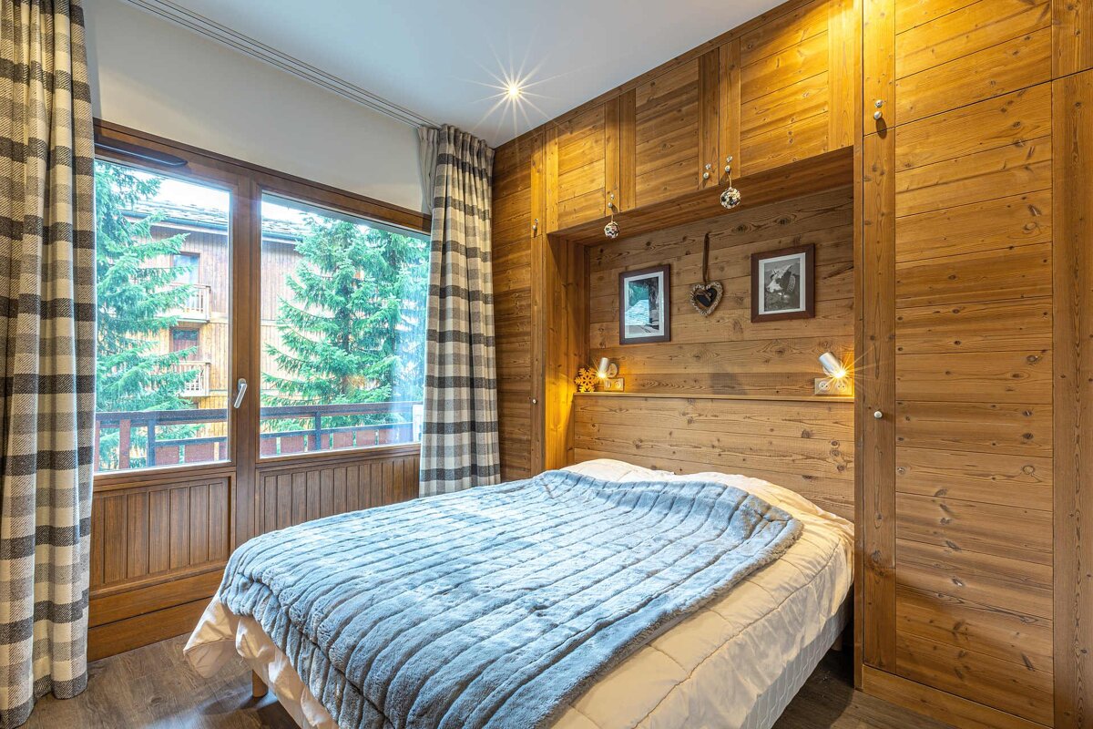 Cozy, wood-paneled bedroom with a comfy bed, light blue blanket, and a large window overlooking evergreen trees.