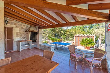 A rustic covered outdoor patio with wooden beams, stone walls, a BBQ, table, chairs, and a view of a garden with a blue ping-pong table.