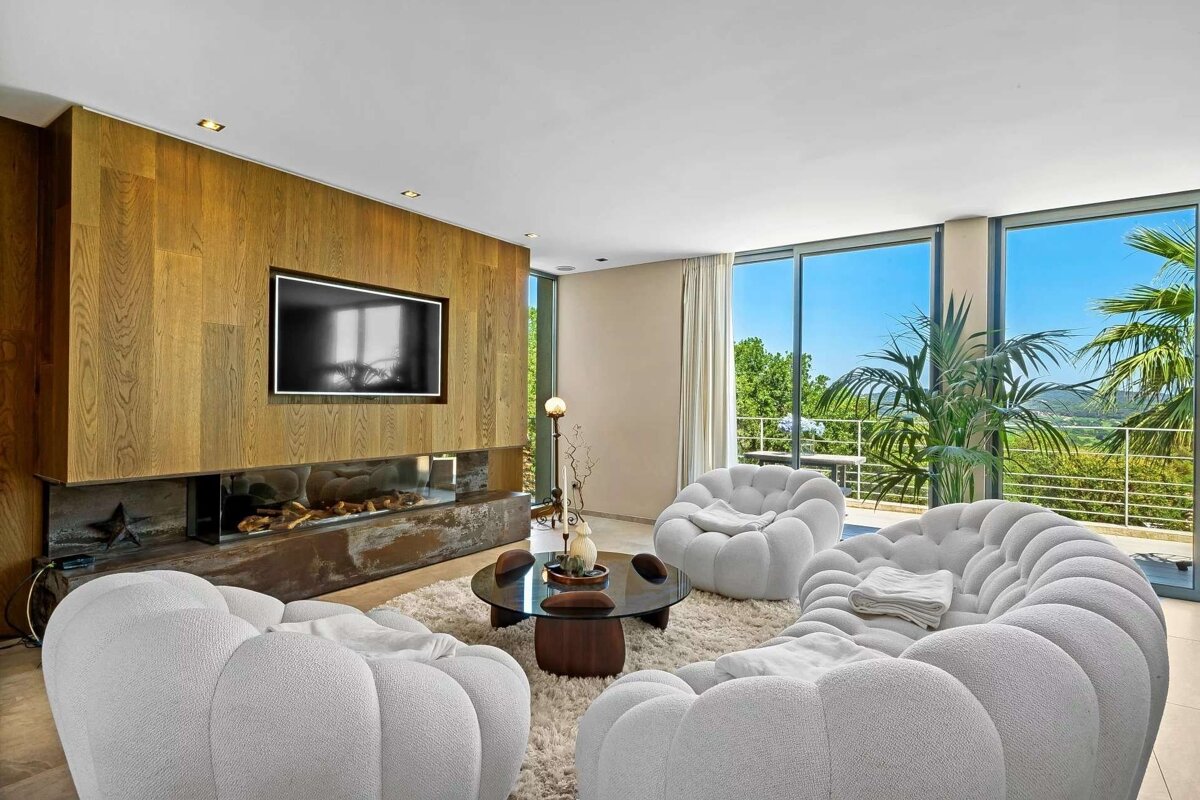 A chic living room with white 'bubble' sofas, a wood-paneled TV wall, a linear fireplace, and floor-to-ceiling windows overlooking a sunny, green landscape.