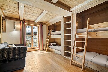 Cozy wooden chalet bedroom featuring built-in bunk beds, wood paneled walls, a sofa, and a door opening to a snowy balcony.