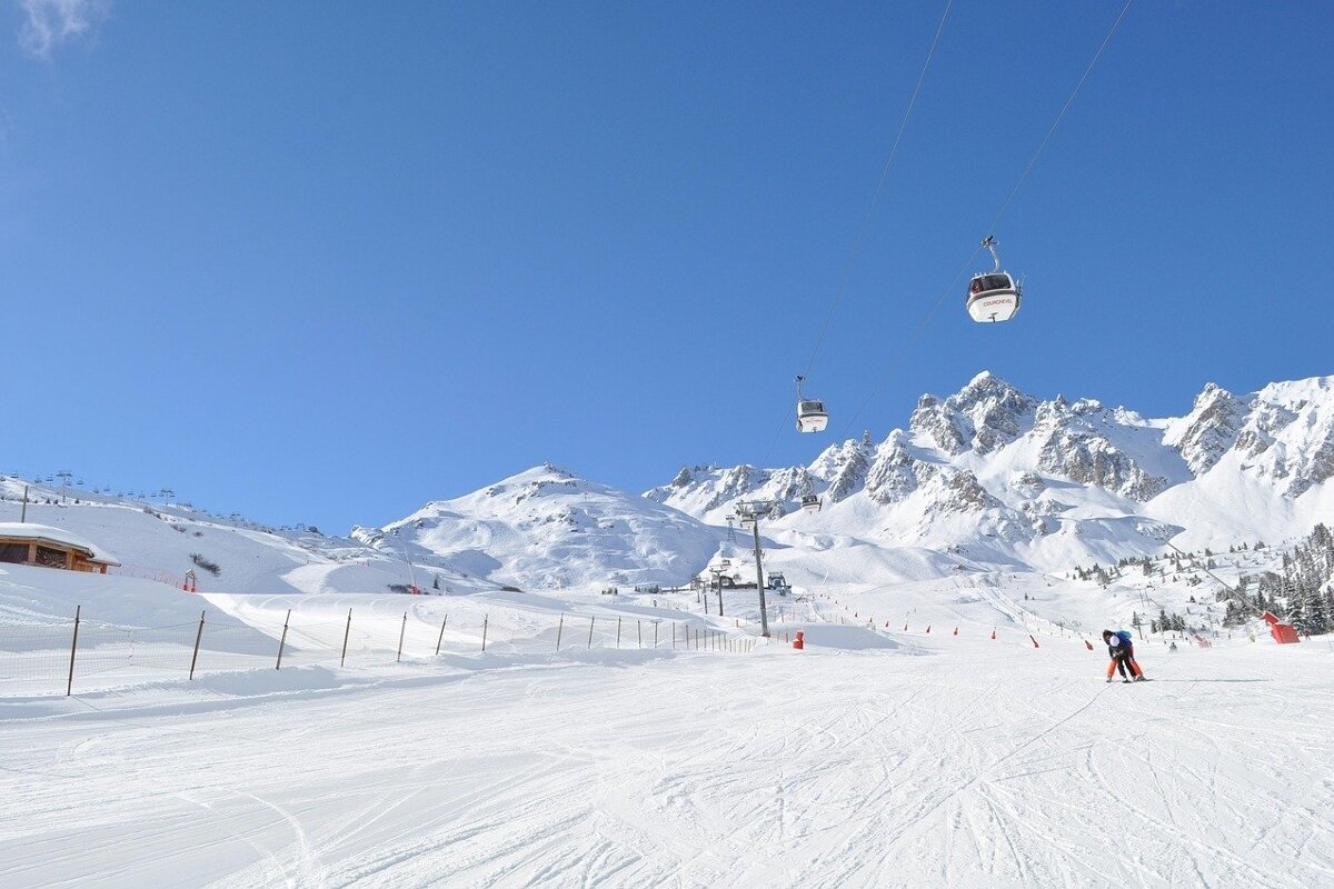 Cable cars glide above a snowy ski slope, with majestic snow-covered mountains under a clear blue sky.