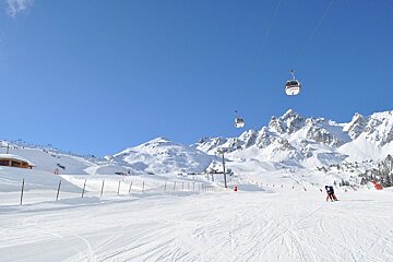 Cable cars glide above a snowy ski slope, with majestic snow-covered mountains under a clear blue sky.
