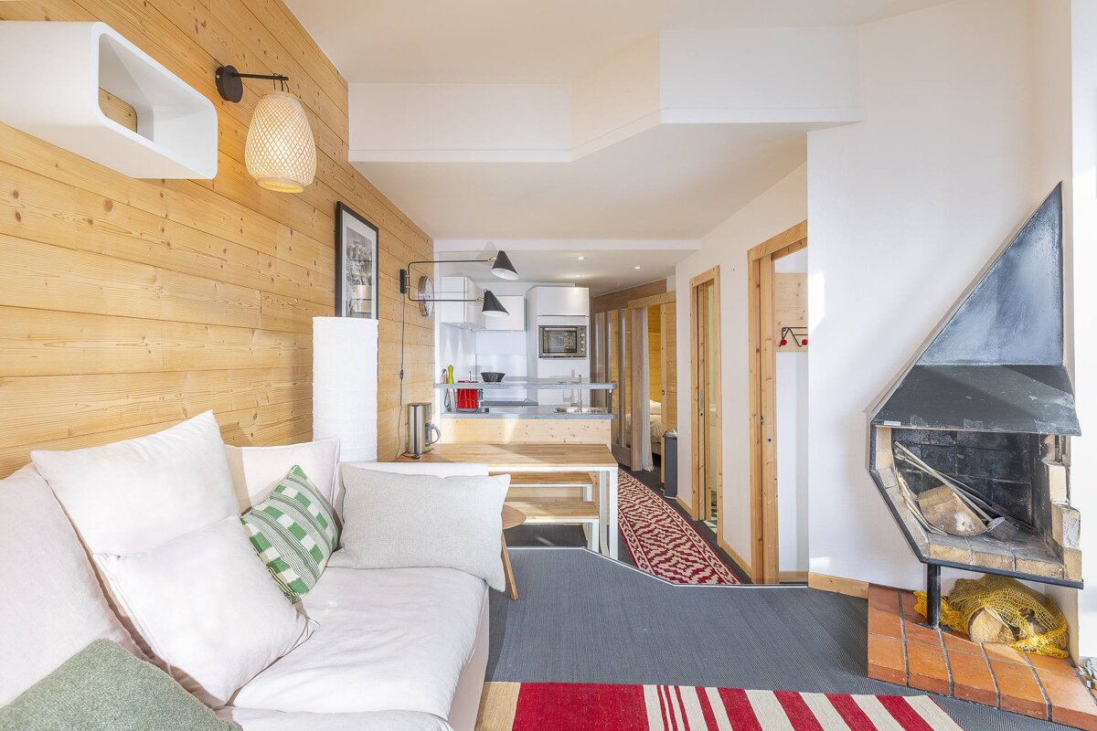 A bright, modern living room featuring a white sofa, wood-paneled wall, and a fireplace, with an open kitchen and hallway visible behind it.