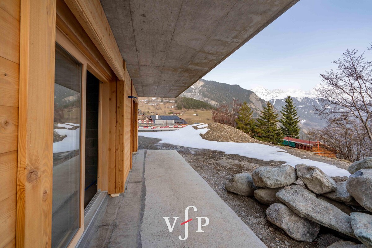 A modern wooden chalet terrace under a concrete overhang, overlooking a scenic snowy mountain landscape.