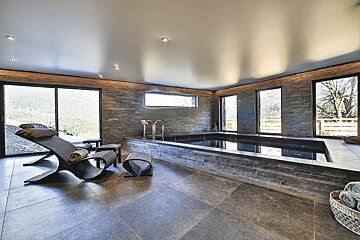 A sleek indoor pool area with stone walls, modern lounge chairs, and large windows revealing a beautiful mountain landscape.