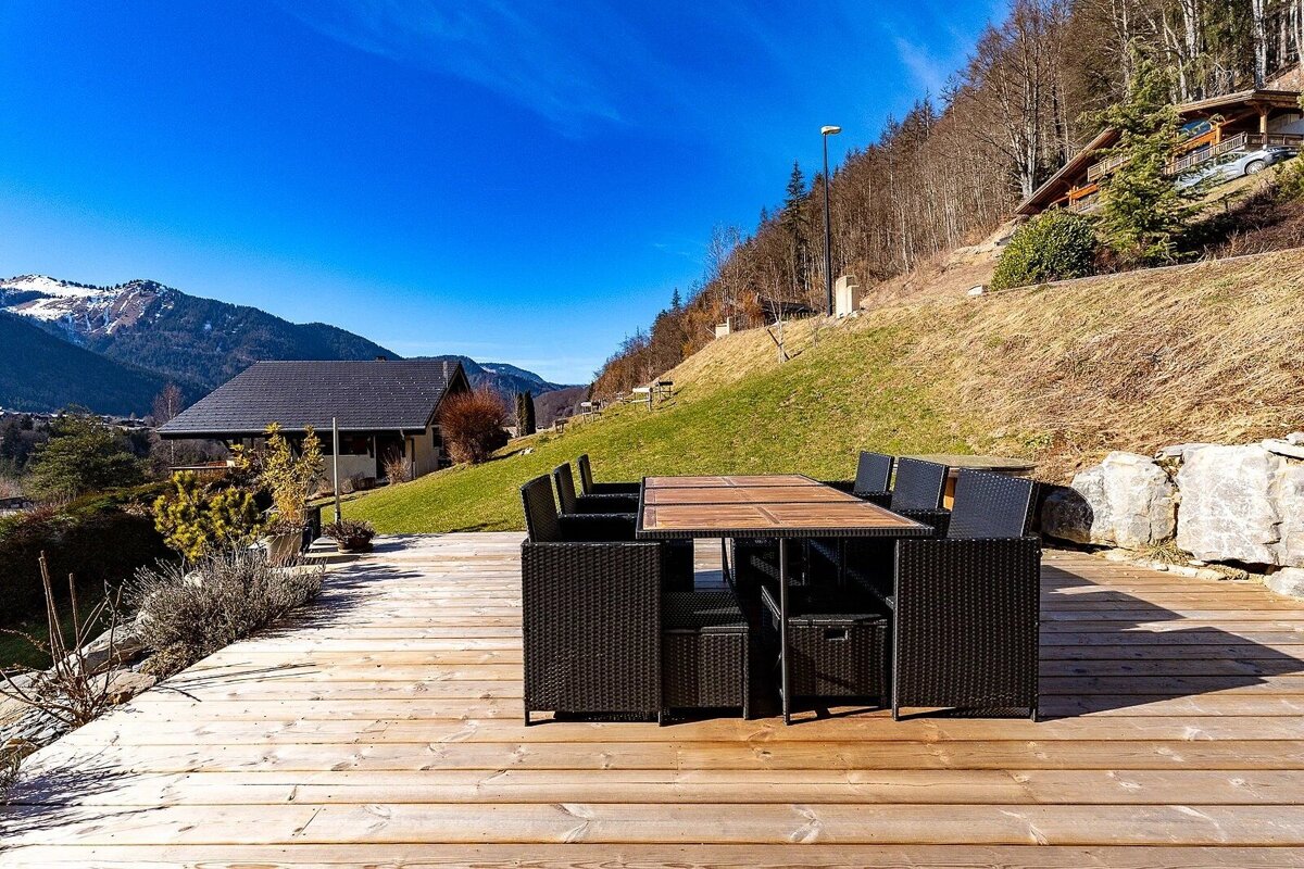 A sunny wooden deck with black outdoor dining furniture, set against a backdrop of green hills, distant snow-capped mountains, and a clear blue sky.