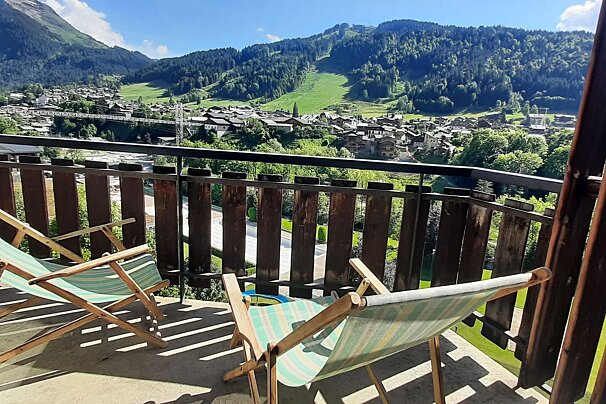 A sunny balcony with two striped deck chairs overlooks a picturesque mountain village nestled among green, forested slopes under a clear blue sky.