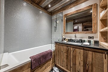 Rustic-modern bathroom featuring grey mosaic tiles, a wooden vanity, a large wood-framed mirror, and a bathtub with a purple towel.