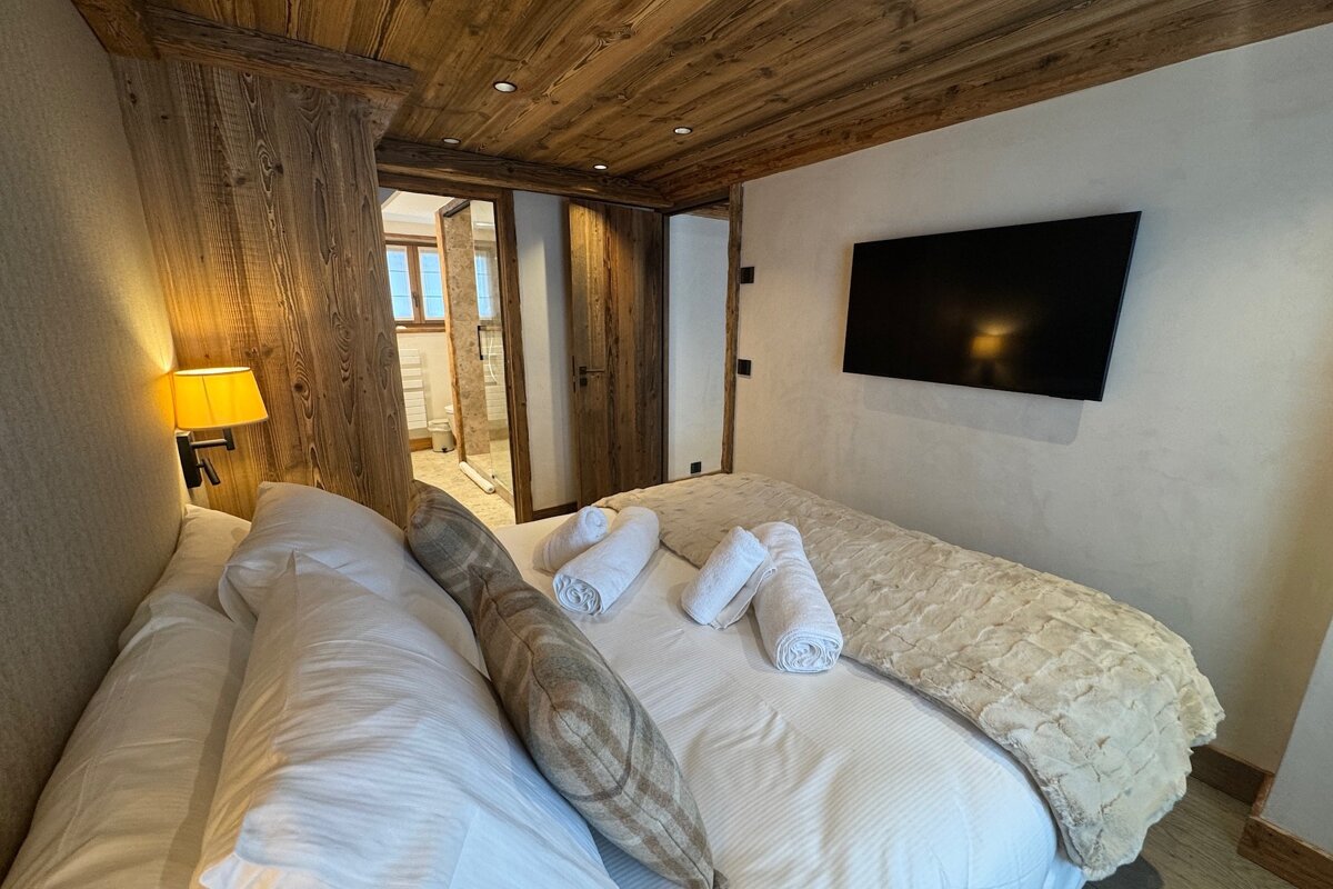Cozy chalet bedroom with wood-paneled ceilings, a neat bed with rolled towels, wall-mounted TV, and an en-suite bathroom.