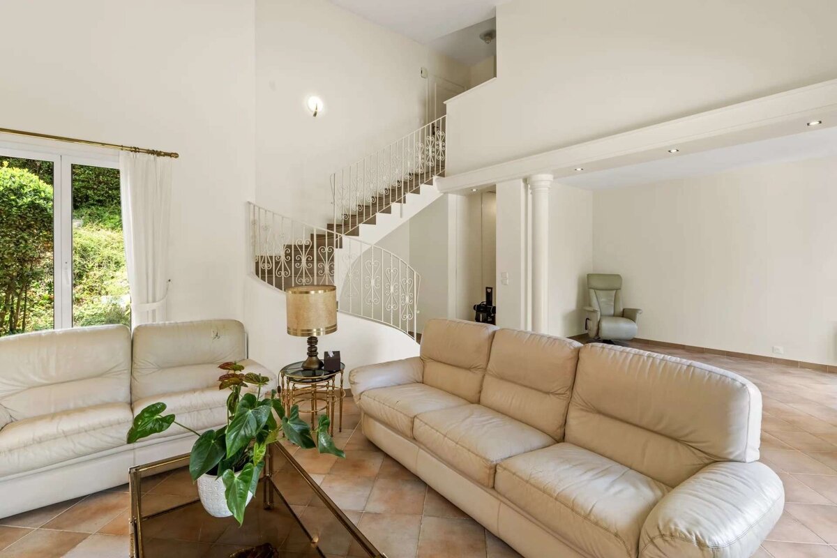 A spacious living room featuring cream leather sofas, a decorative white spiral staircase, large windows looking out to greenery, and terracotta tiled floors.