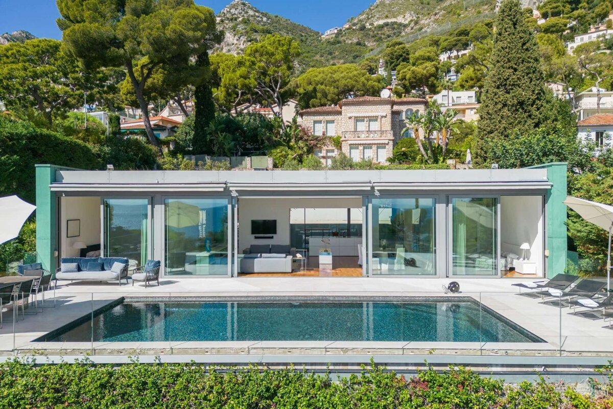 A modern house with a pool and panoramic glass walls overlooks a lush, mountainous landscape with distant sea views on a sunny day.
