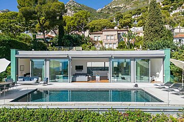 A modern house with a pool and panoramic glass walls overlooks a lush, mountainous landscape with distant sea views on a sunny day.