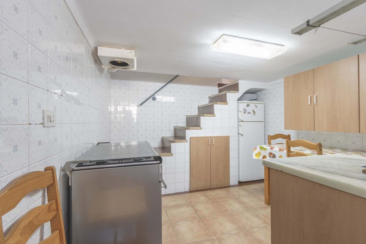 A small, tiled kitchen features wooden cabinets, appliances, a dining area, and a unique staircase built into the space leading upstairs.