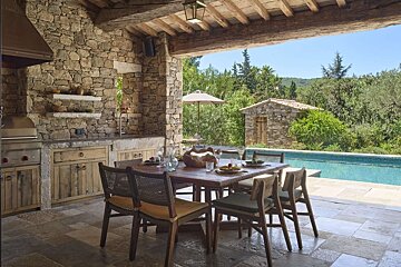 Rustic outdoor patio with stone walls, wooden beams, a dining table, and outdoor kitchen, overlooking a pool and lush landscape under a blue sky.