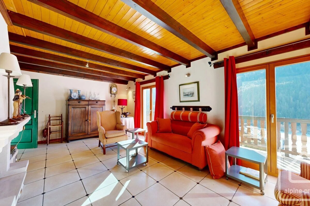 Rustic chalet living room with a wooden beamed ceiling, red sofa, armchair, tiled floor, and glass doors opening to a scenic balcony.