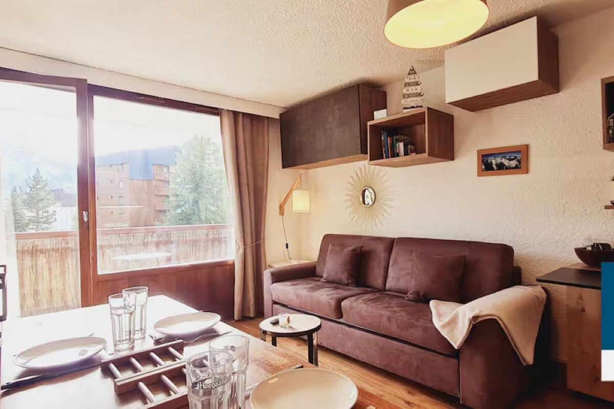 A cozy living room features a brown sofa, a wooden dining table set for two, and a bright balcony with mountain and tree views. Floating shelves decorate the wall.
