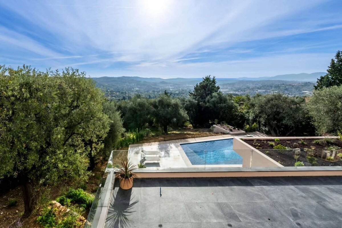 A luxurious modern patio with a bright blue infinity pool and glass railing, offering stunning panoramic views of a lush green valley under a sunny sky.