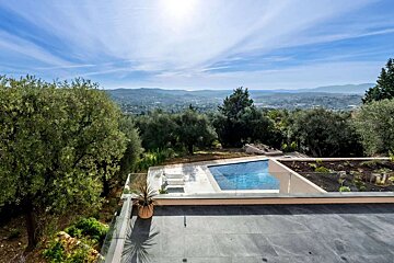 A stunning outdoor patio with an infinity pool offering panoramic views of a lush valley, distant town, and rolling hills under a sunny blue sky.