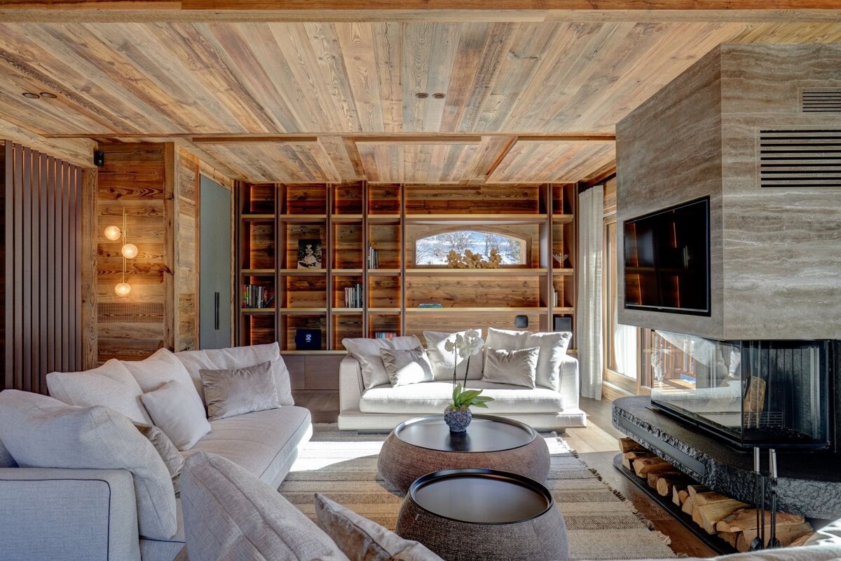 Luxurious chalet living room with extensive wood paneling, plush light sofas, a modern fireplace, built-in shelves, and a snowy view.