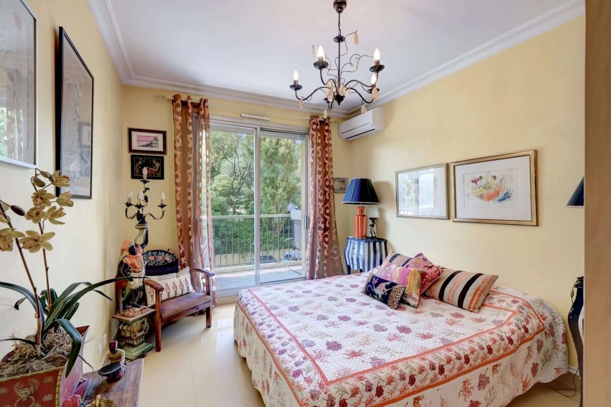A bright, eclectic bedroom with yellow walls, a patterned bedspread, chandelier, and sliding glass doors leading to a balcony with lush views.