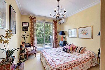 A bright, eclectic bedroom with yellow walls, a patterned bedspread, chandelier, and sliding glass doors leading to a balcony with lush views.