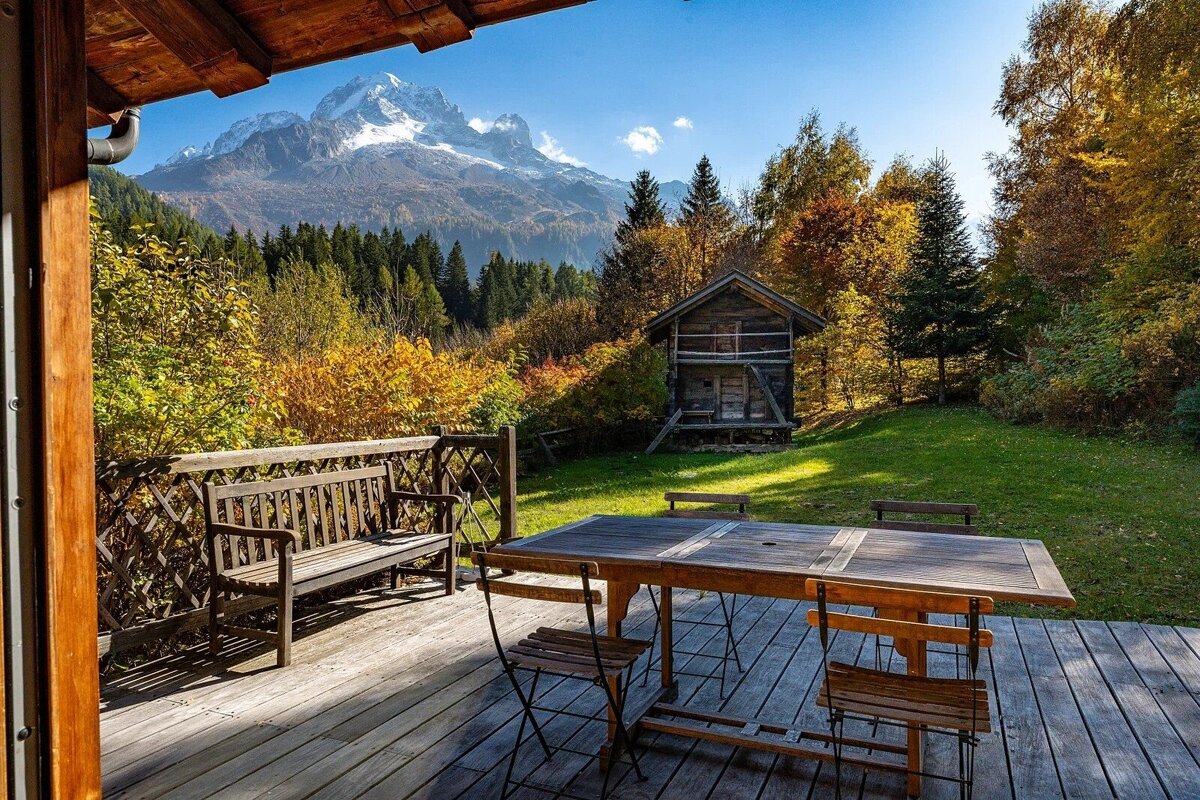 View from a wooden deck: autumn trees, a small cabin, and majestic snow-capped mountains under a clear blue sky.
