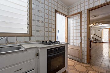 A dated kitchen with patterned tiled walls and floor, featuring a sink, stove, and oven. An open door reveals a traditional living space with stairs.