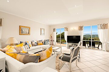 A bright, spacious living room with white sofas, yellow accent cushions, and sliding glass doors opening to a balcony with scenic views.