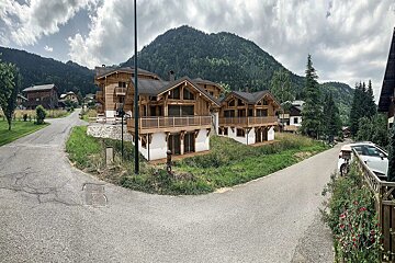 Wooden mountain chalets nestled between winding roads, with lush green peaks in the background under a cloudy sky.
