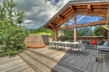 A modern wooden chalet with large glass windows and an outdoor deck featuring a dining table and hot tub, set against a green mountain landscape under a cloudy sky.