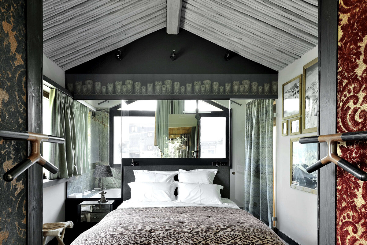 A cozy, eclectic bedroom with a slanted wooden ceiling, a patterned duvet, a large mirror over the bed, and an owl motif band.