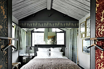 A cozy, eclectic bedroom with a slanted wooden ceiling, a patterned duvet, a large mirror over the bed, and an owl motif band.
