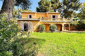 A sunny, yellow Mediterranean-style house with a balcony, arched porch, and purple shutters, nestled amidst green grass and trees.