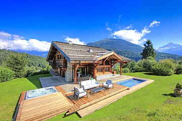 A luxurious wooden chalet in lush green mountains features a hot tub, swimming pool, and outdoor patio furniture under a bright blue sky.