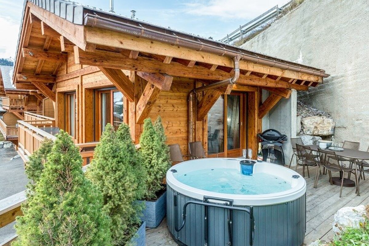 Cozy wooden chalet deck featuring a round outdoor hot tub, potted evergreen shrubs, and patio dining furniture.