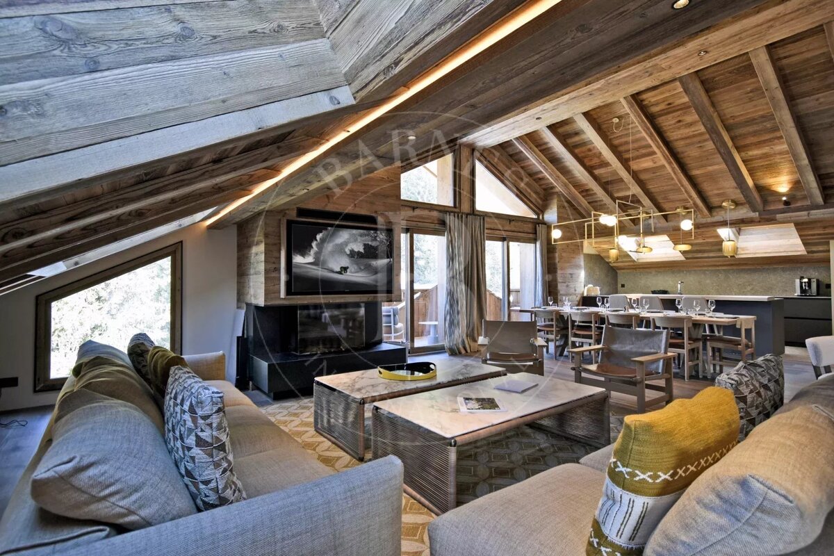 Luxurious, open-plan chalet living room with vaulted wooden ceilings, a large TV above a fireplace, cozy sofas, and a dining area.