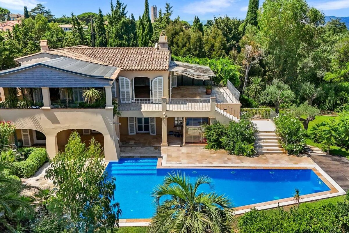 Luxurious multi-story villa with terracotta roof, balconies, and a vibrant blue swimming pool, nestled among lush green trees and gardens.