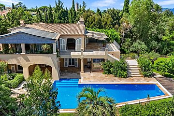 Luxurious multi-story villa with terracotta roof, balconies, and a vibrant blue swimming pool, nestled among lush green trees and gardens.