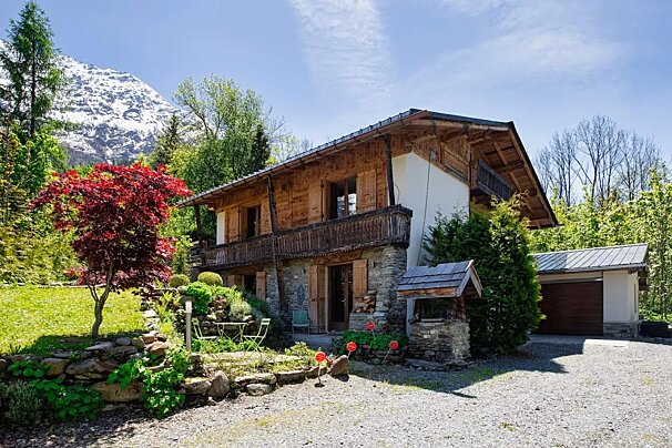 A rustic wooden chalet surrounded by lush greenery and a vibrant red tree, with snow-capped mountains in the background.